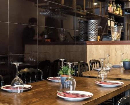 Restaurant dining room with a set wooden table, black chairs, dark tiled walls, glassware, and plants.