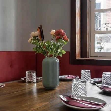 Dining table with a green vase of red and white flowers, place settings, red seating, and a window.