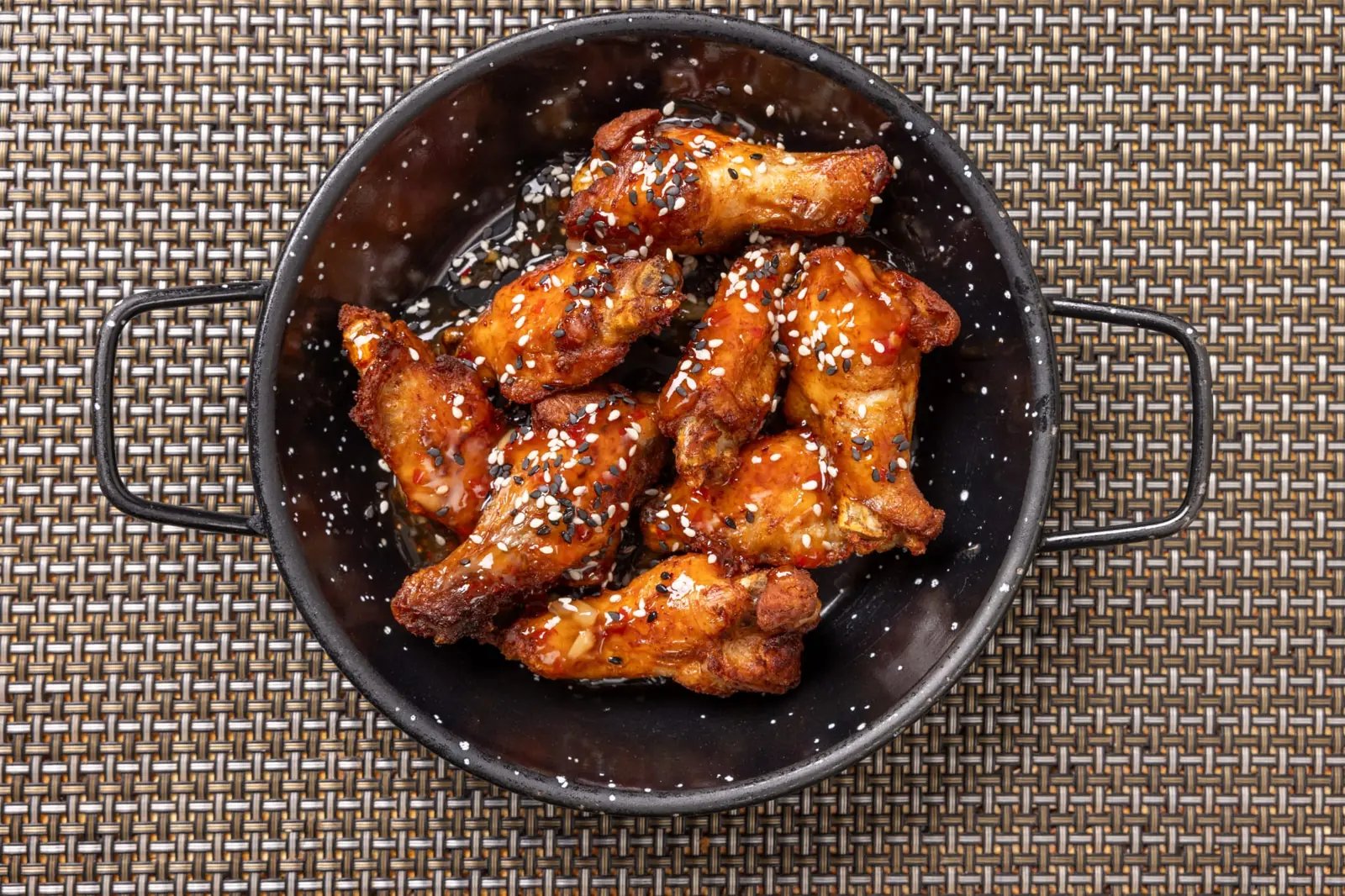 Glazed sesame-topped fried chicken wings in a black handled bowl on a woven placemat.