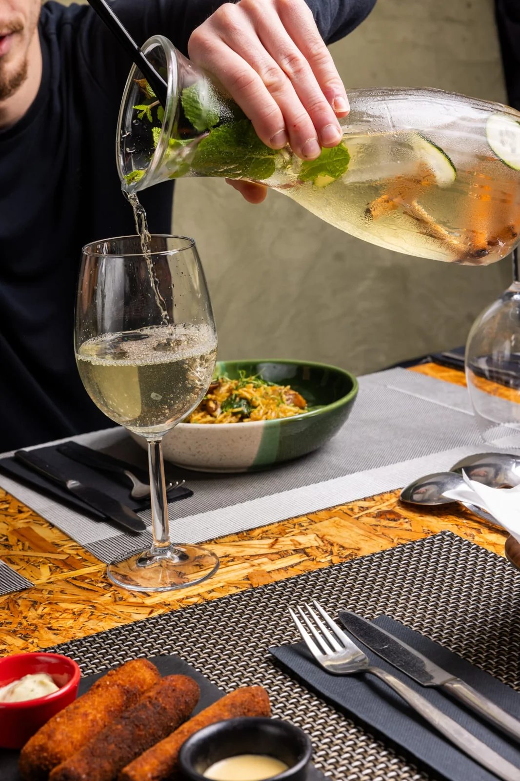 Person pouring a pale sparkling drink with mint and cucumber into a wine glass at a dining table.