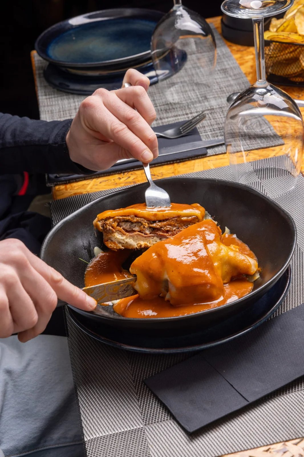 Person cutting a layered dish covered in orange sauce on a black plate at a table.