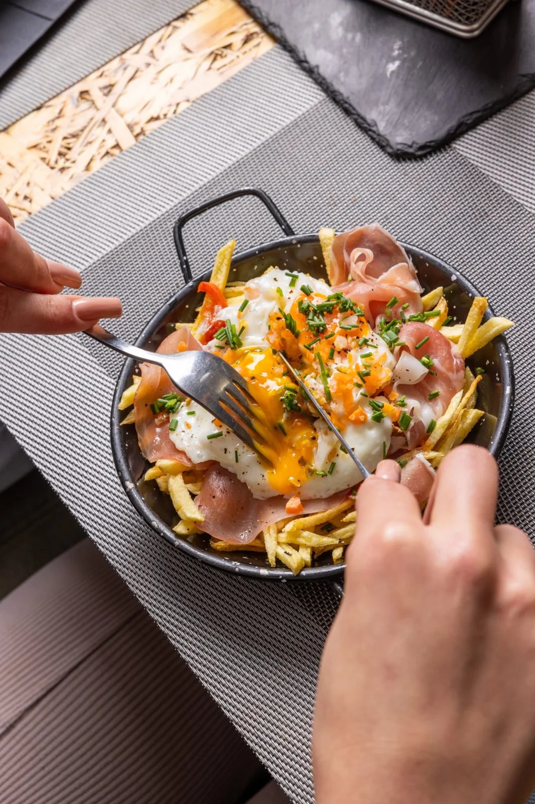 Skillet of fries topped with prosciutto, fried eggs, chives, and vegetables, being cut with fork and knife.